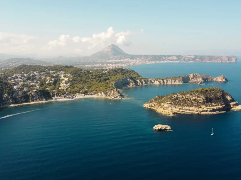 Luchtpanorama van Jávea's kust: weelderige heuvels met residenties, een zandstrand en een rotsachtig eiland in de helderblauwe zee. Een zeilboot en het kielzog van een boot zijn zichtbaar, met de Montgó-berg aan de horizon.