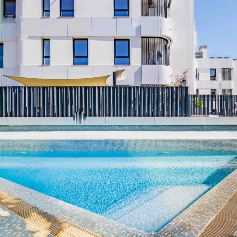 Gemeenschappelijk zwembad met blauwe mozaïektegels en inlooptrap, gelegen bij een modern wit appartementencomplex met strakke omheining en mediterrane beplanting.