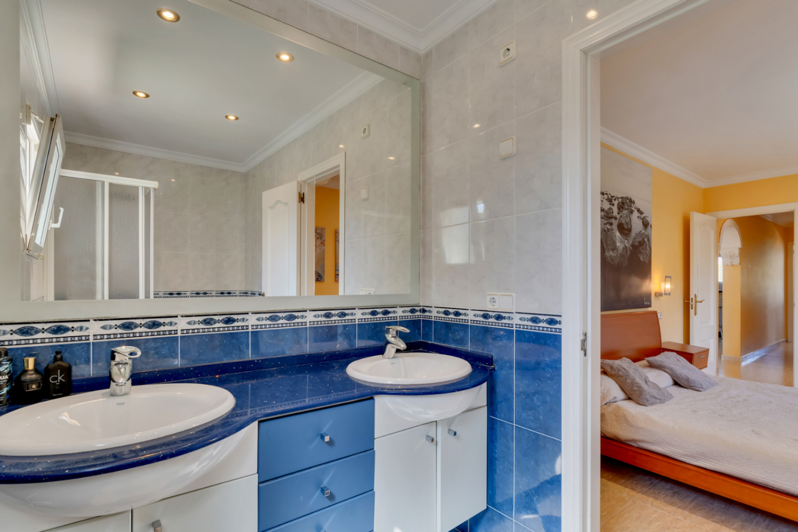 Badkamer met dubbele wastafel op een blauw blad, decoratieve betegeling en een grote spiegel. Directe verbinding met de slaapkamer in warme tinten.