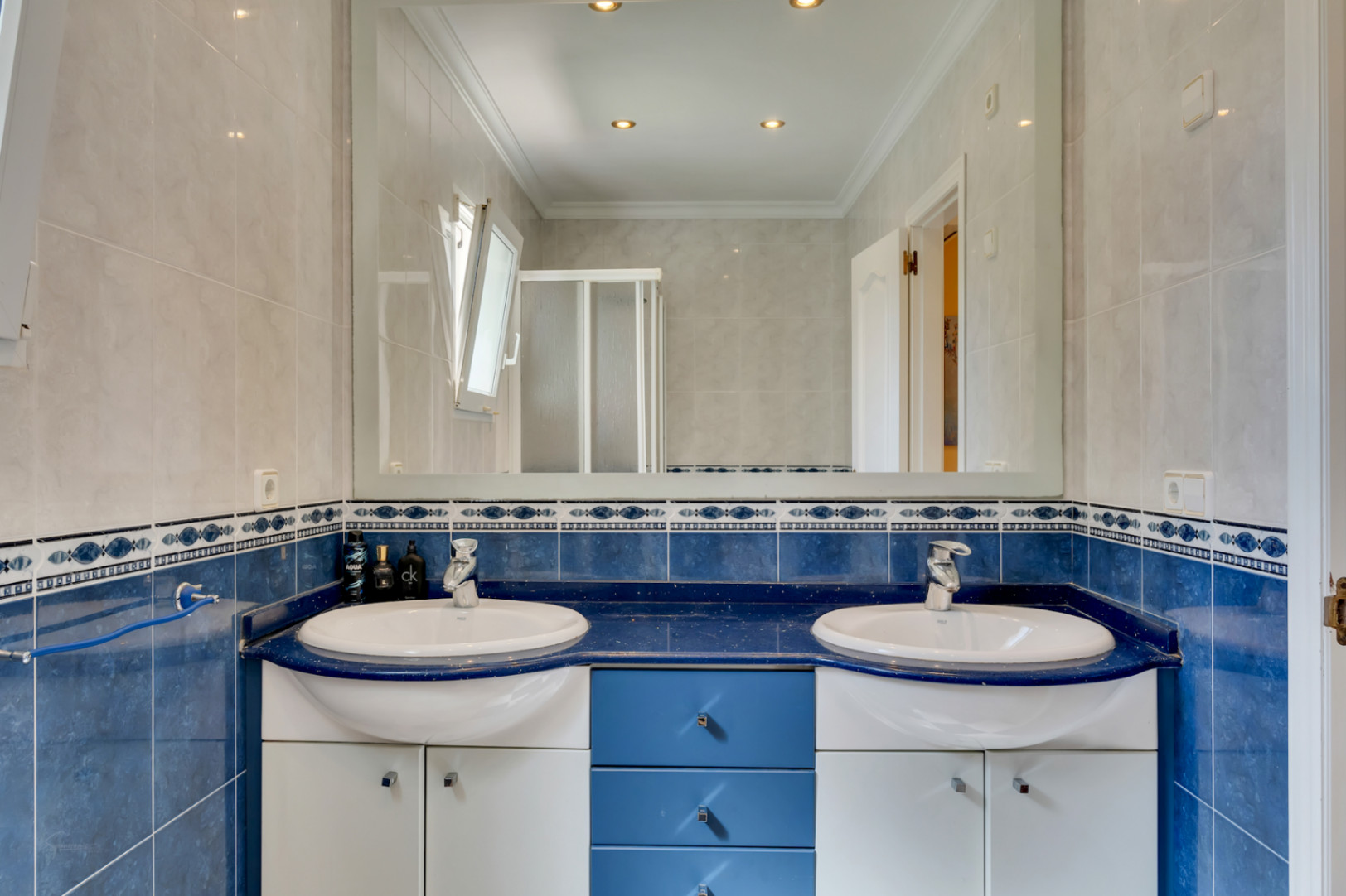 Badkamer met dubbele wastafel op een blauw werkblad, grote wandspiegel, inbouwspots en tweekleurige wandtegels met een decoratieve sierrand.