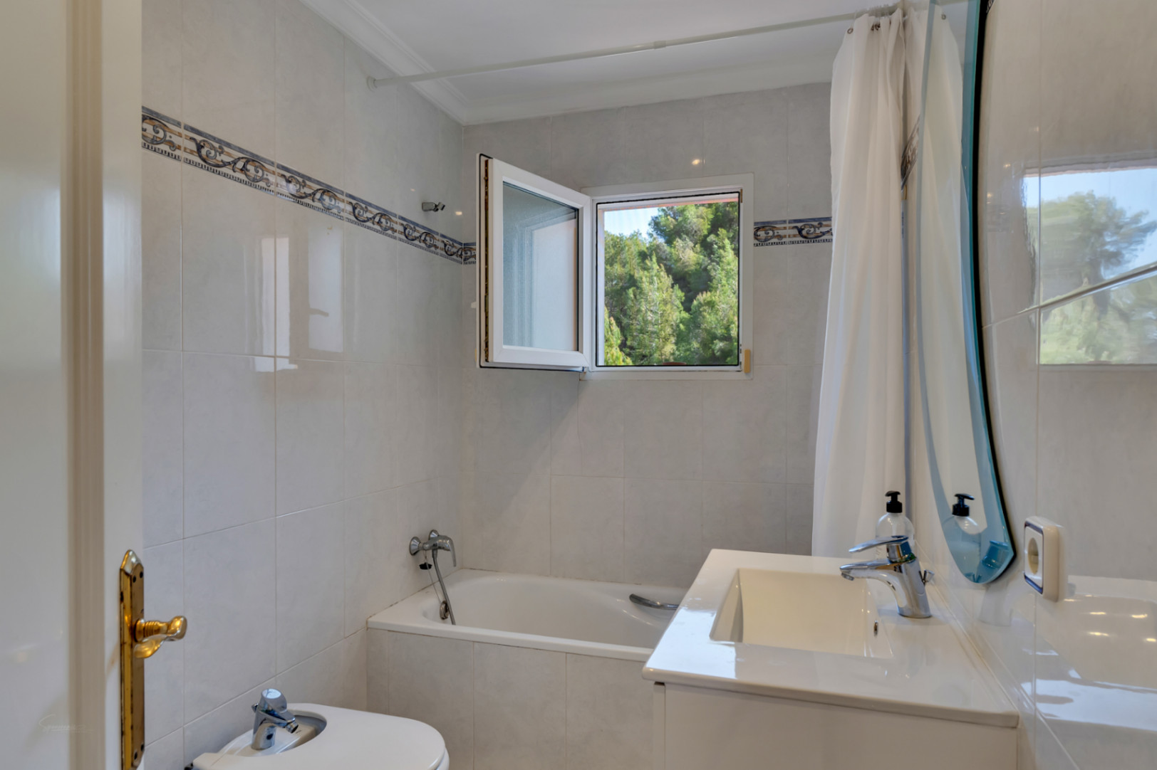 Functionele badkamer met ligbad, bidet en wit wastafelmeubel. Een draaikiepraam biedt natuurlijk licht en uitzicht op de omliggende groene vegetatie.