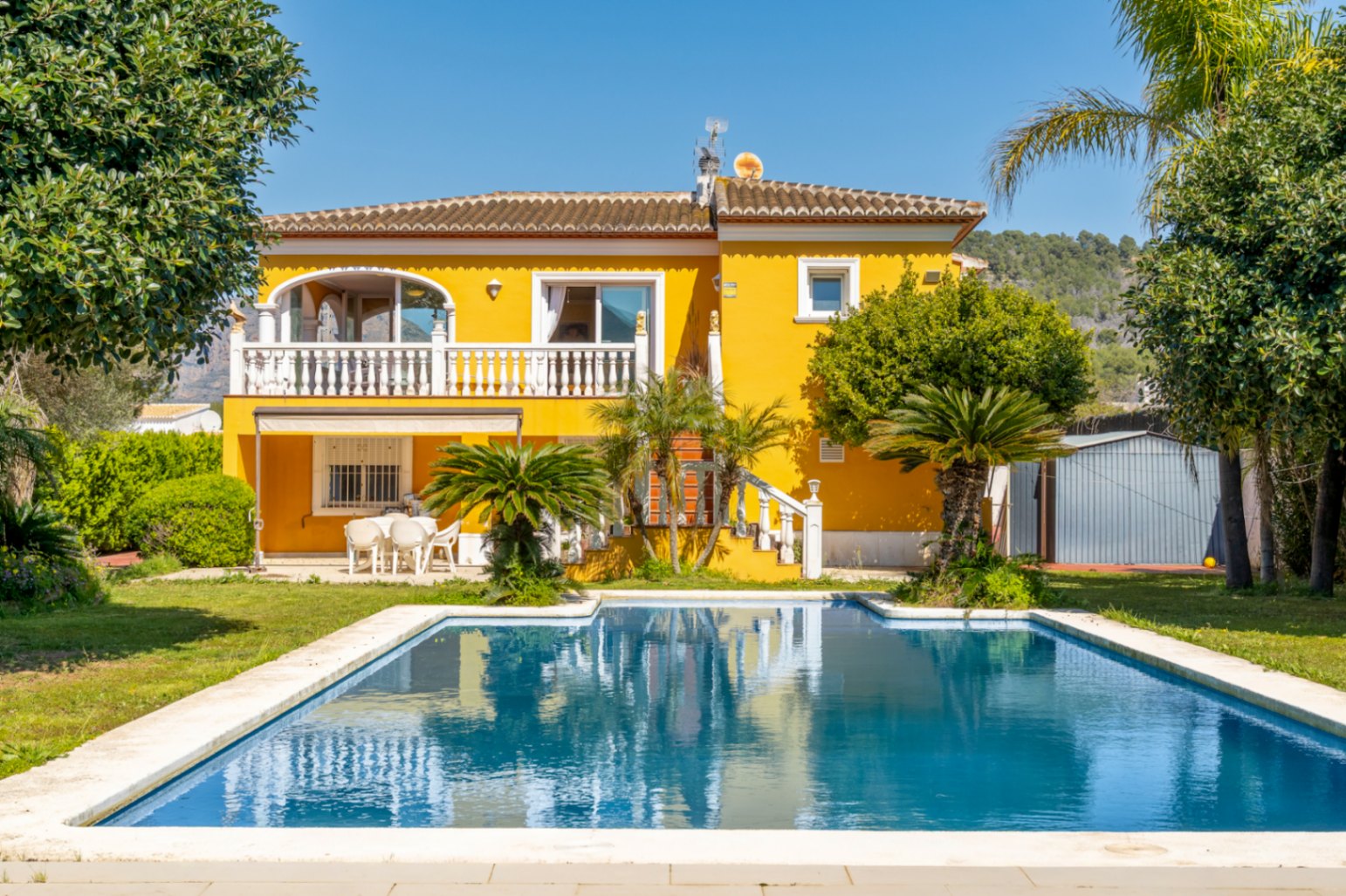 Gele villa-gevel met een groot rechthoekig zwembad, boventerras met witte balustrades en een mediterrane tuin met palmbomen.