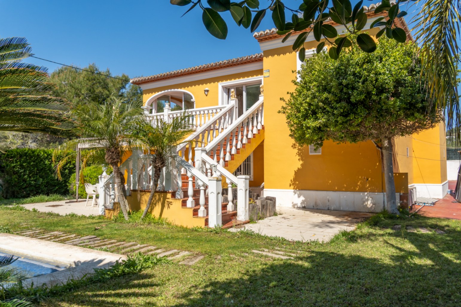 Gele villa met een buitentrap voorzien van witte balustrades die naar het boventerras leidt. Mediterrane tuin met palmbomen, gazon en zicht op de rand van het zwembad.
