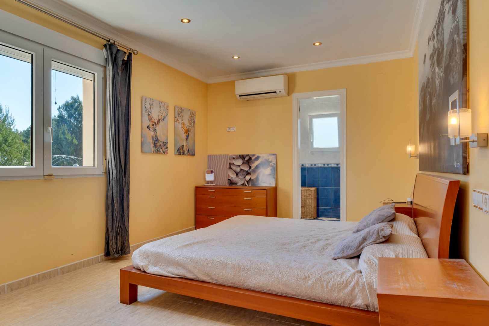 Lichte slaapkamer met en-suite badkamer, airconditioning en een groot PVC-raam. Voorzien van een tegelvloer, houten meubilair en inbouwspots in het plafond.