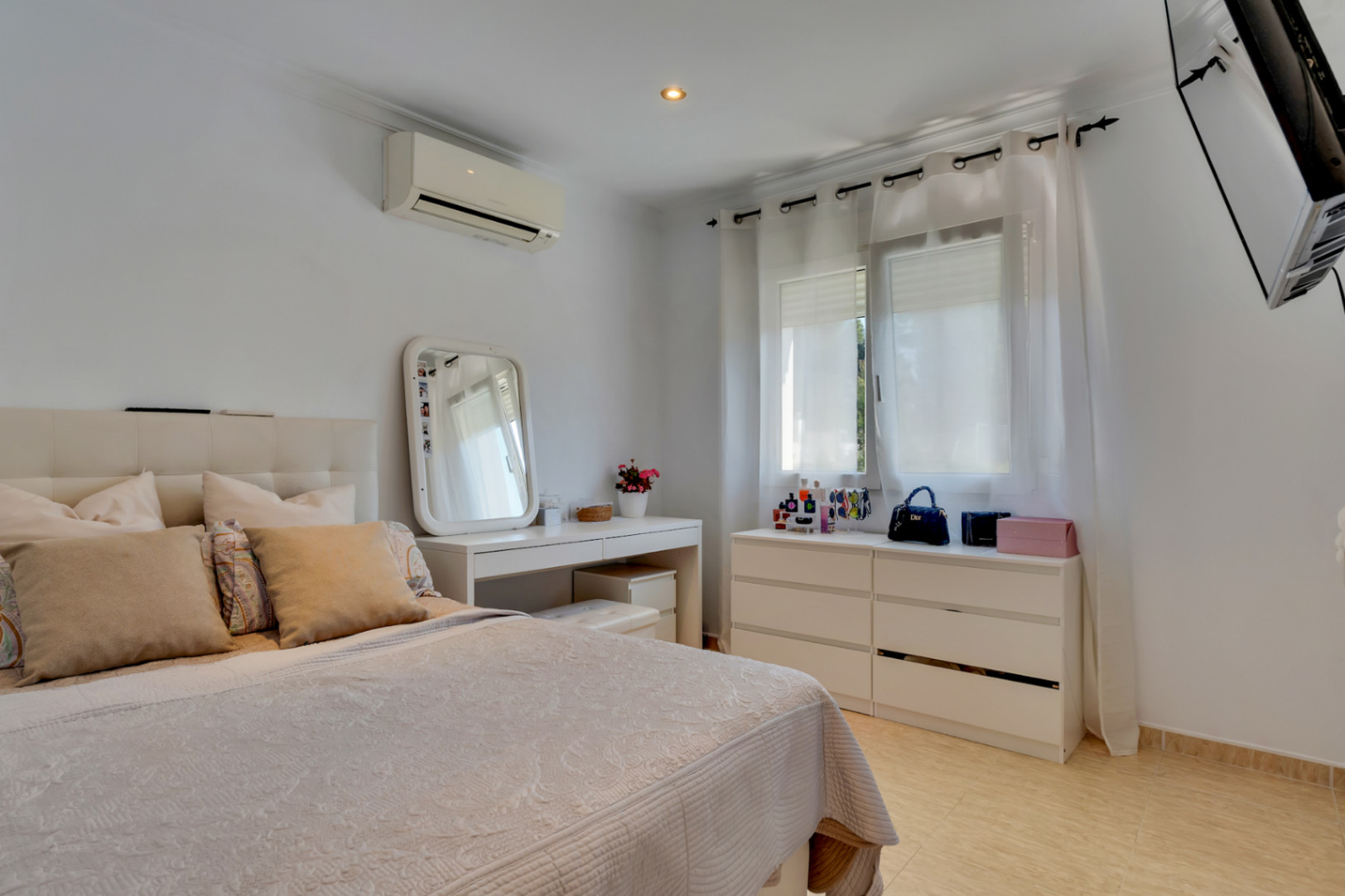 Lichte slaapkamer met wit meubilair, een kaptafel, airconditioning en een tegelvloer. Het raam met vitrage laat zacht natuurlijk licht binnen in de ruimte.