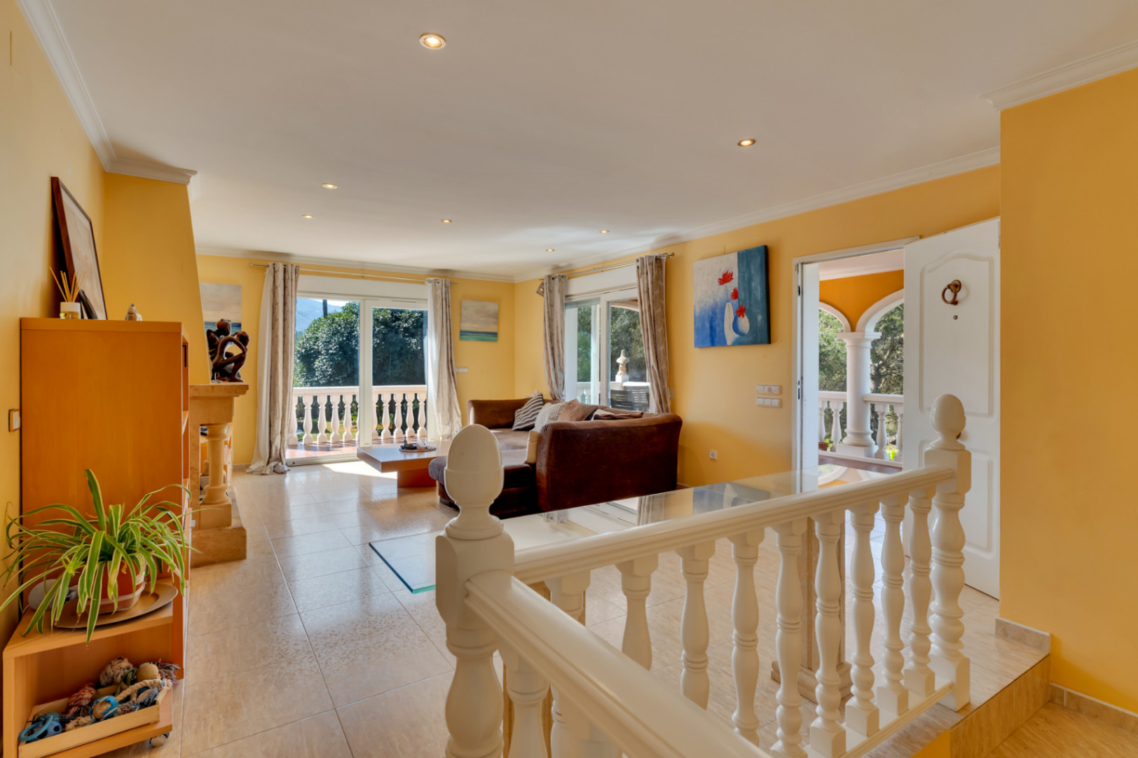 Lichte woonkamer met tegelvloer en okergele wanden, verbonden met een terras via grote schuifdeuren. De witte balustrade markeert de overgang naar de zithoek.