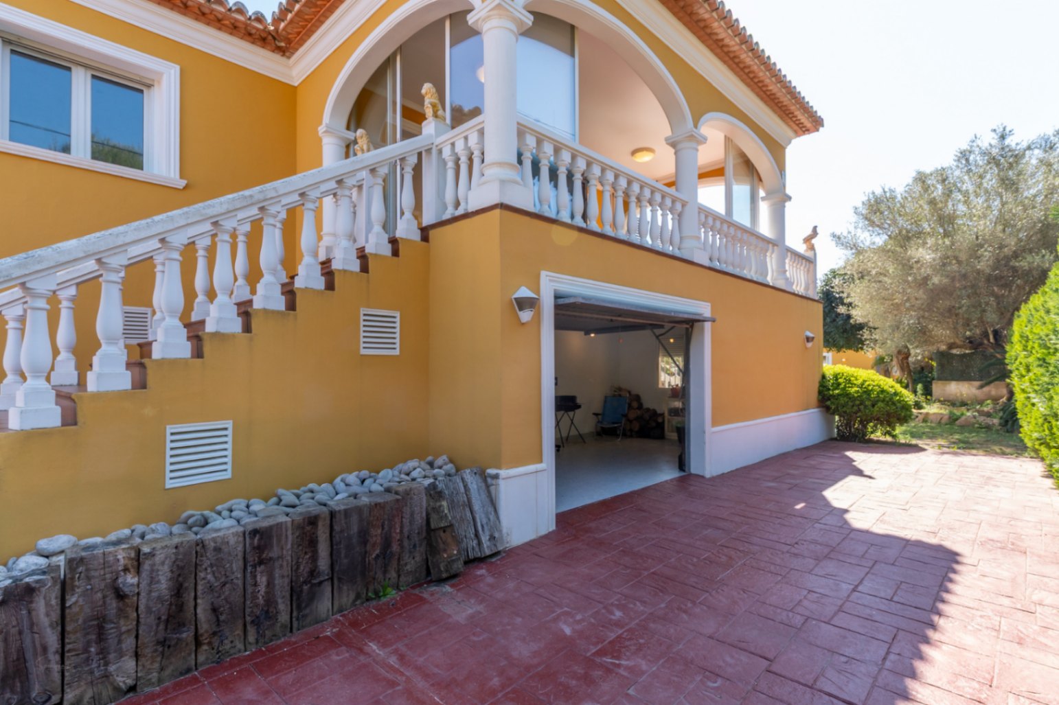 Mediterrane gevel met een buitentrap en witte balustrade die leidt naar een overdekt terras met bogen boven een inpandige garage en bestraat terras.