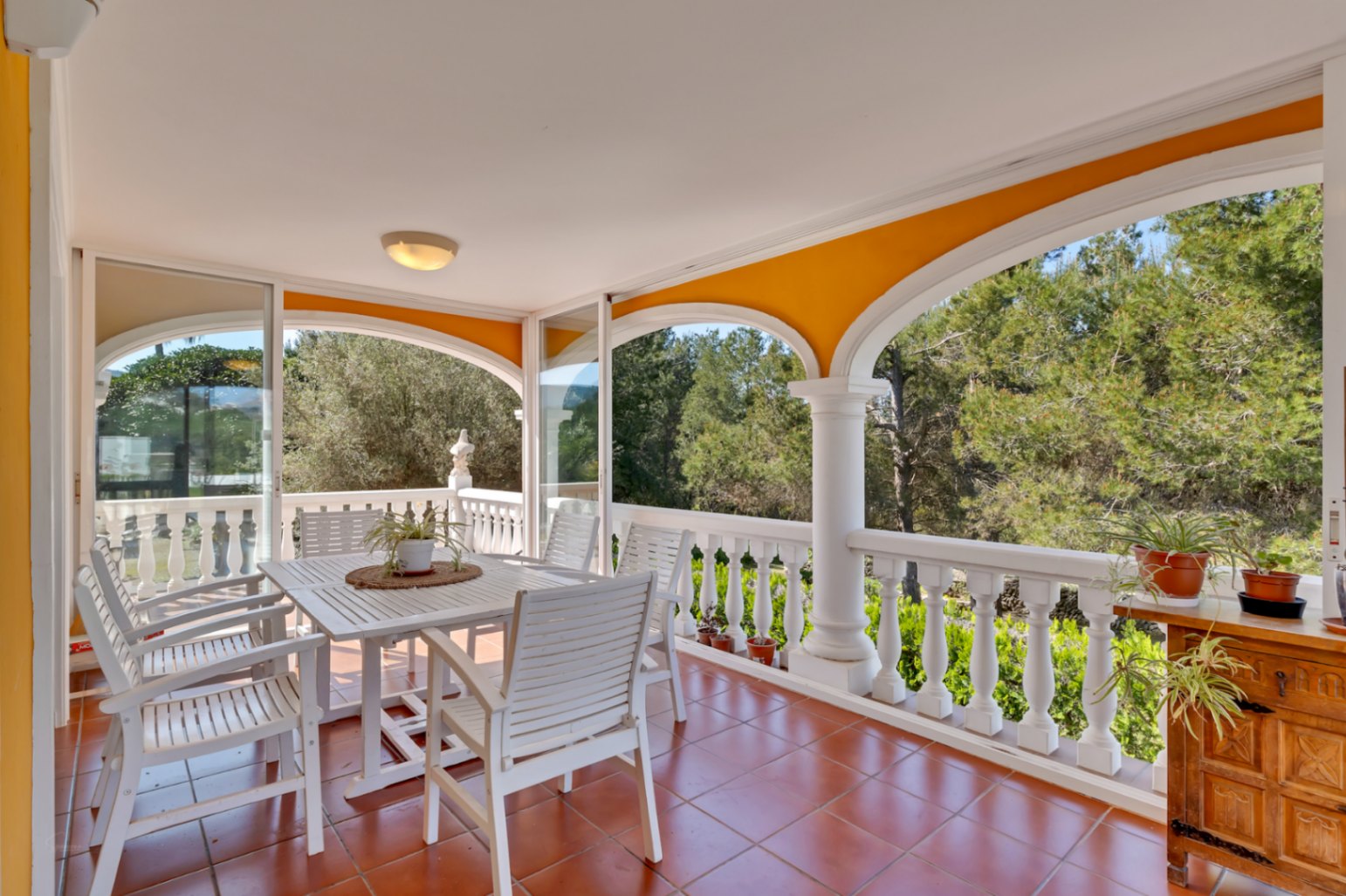 Overdekt terras met witte bogen, een klassieke balustrade en terracotta tegels. Voorzien van een witte houten eethoek en vrij uitzicht op de omliggende mediterrane natuur.