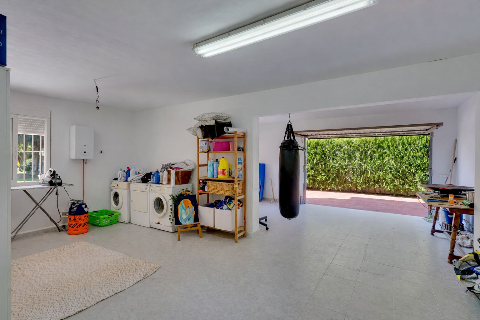 Ruime multifunctionele ruimte met een wasruimte, opbergrekken en een fitnesshoek met bokszak, die direct toegang biedt tot de oprit via de garagedeur.