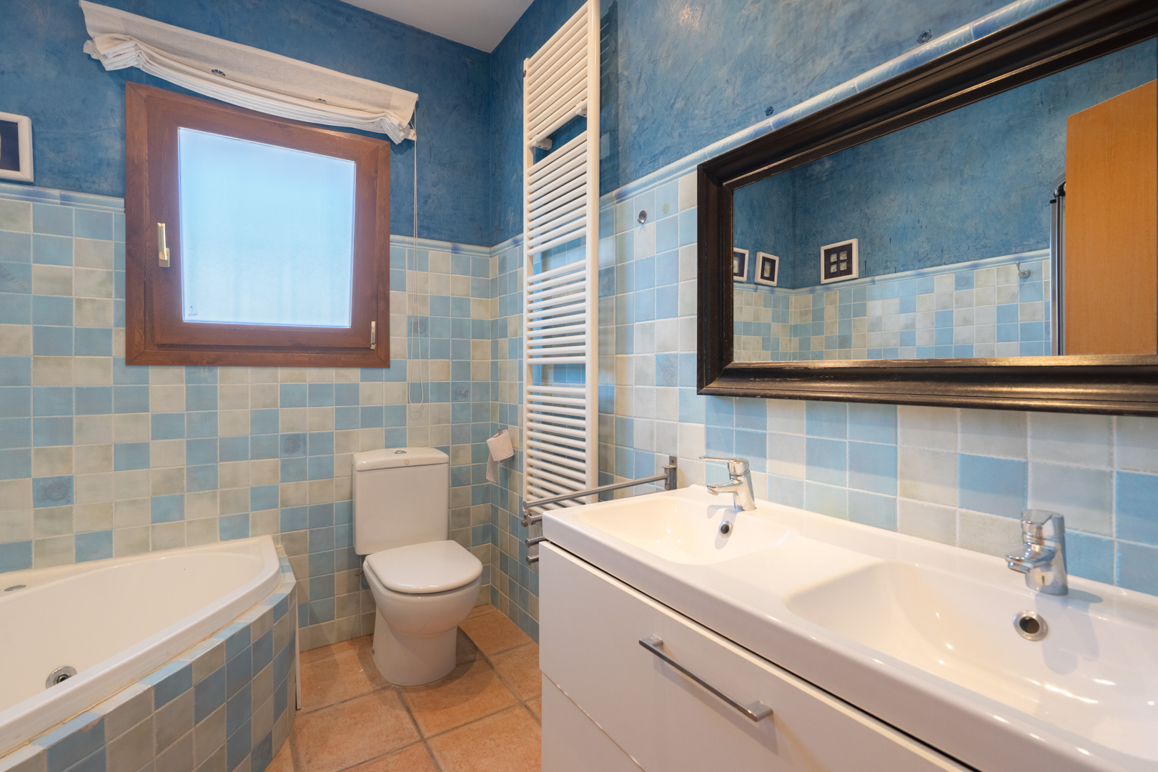Badkamer met blauw geblokte wandtegels, een dubbel wastafelmeubel, hoekbad en witte handdoekradiator gecombineerd met blauw stucwerk op de muren.