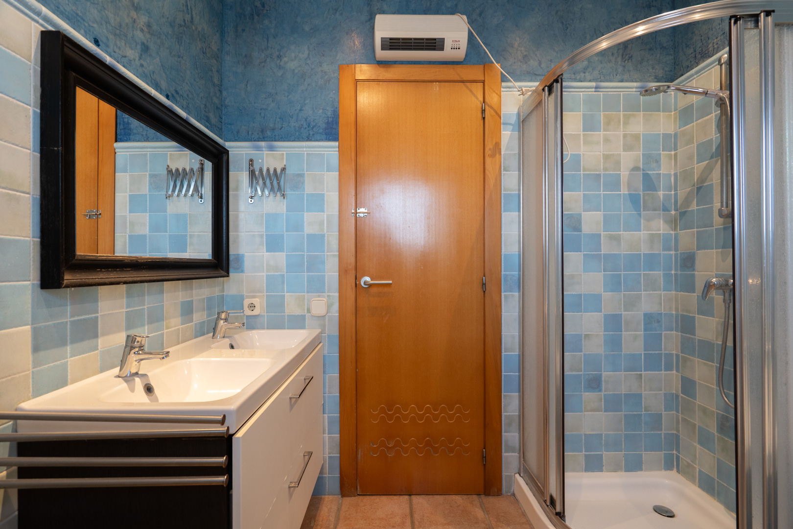 Badkamer voorzien van een dubbele wastafel, glazen hoekdouche en wanden met blauwe mozaïektegels en een decoratieve houten deur.
