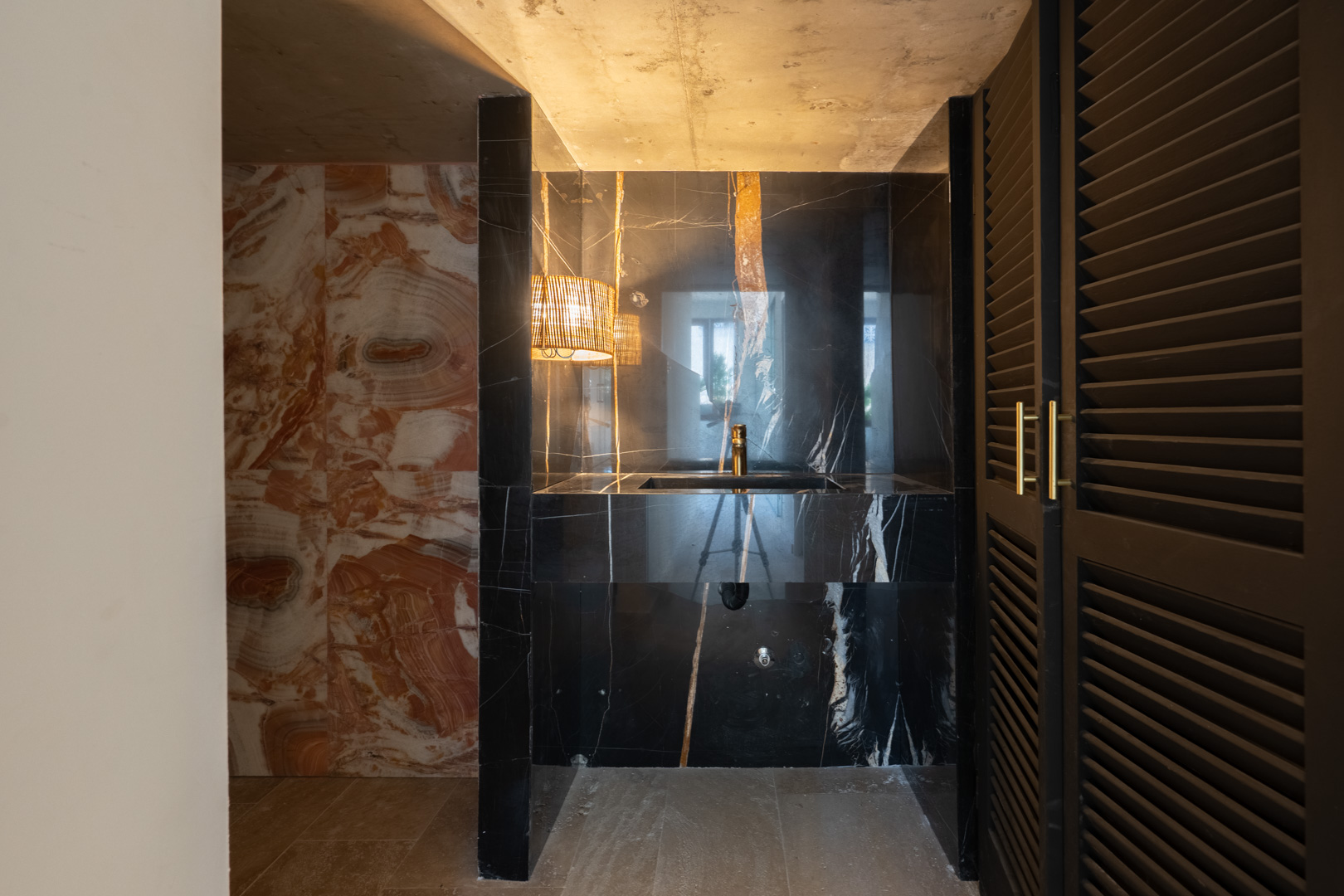 Design toiletruimte met een geïntegreerde wastafel van zwart geaderd marmer, gouden kranen, natuurstenen wanden en donkere louvredeuren met goudkleurig beslag.