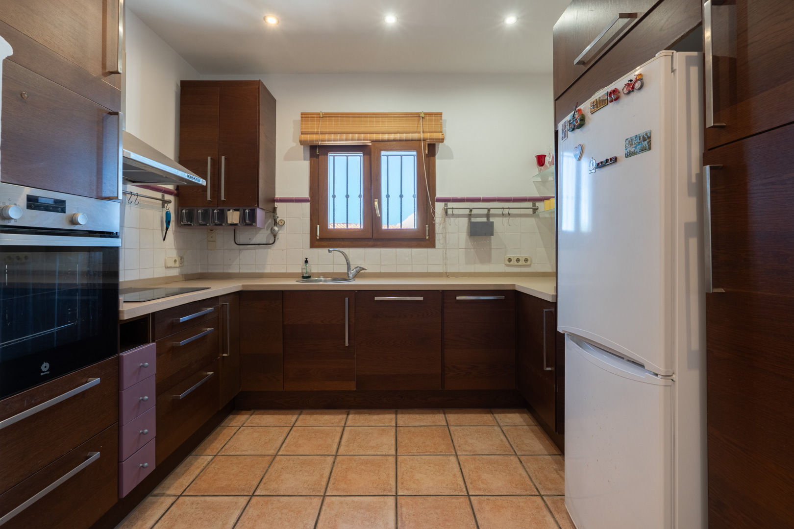 Functionele keuken met donkerhouten kasten, beige aanrechtblad en terracotta vloertegels. Voorzien van een inbouwoven, witte koelkast en inbouwspots.