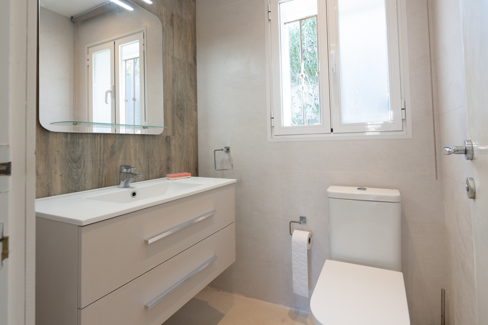 Moderne badkamer met hangend wastafelmeubel, houten accentmuur en wit toilet. Voorzien van een raam voor natuurlijke ventilatie en lichtinval.