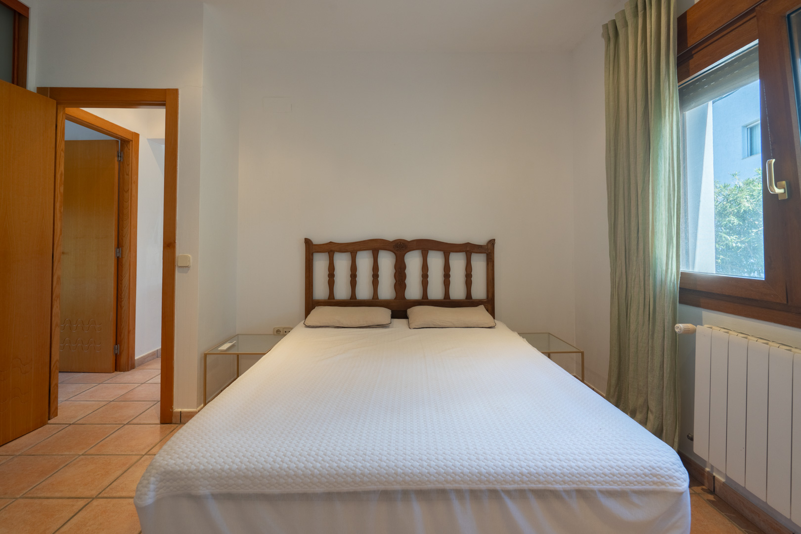 Slaapkamer met tweepersoonsbed, bewerkt houten hoofdbord en glazen nachtkastjes. De ruimte is voorzien van een tegelvloer, radiator en groene gordijnen.