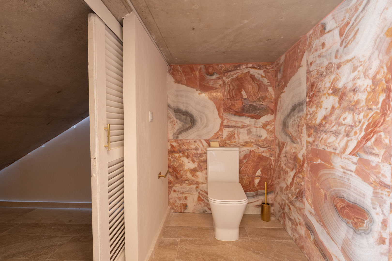 Toiletruimte met roze geaderde marmeren wandbekleding, modern wit toilet en zichtbeton plafond. Voorzien van een witte lamellen schuifdeur en goudkleurige accenten.