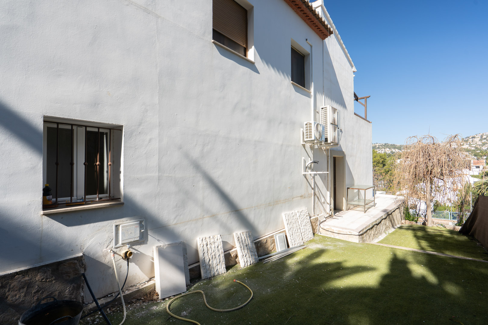 Witte zijgevel met natuurstenen plint, externe airconditioningunits en een tuinzone met kunstgras en vrij uitzicht op de omgeving.