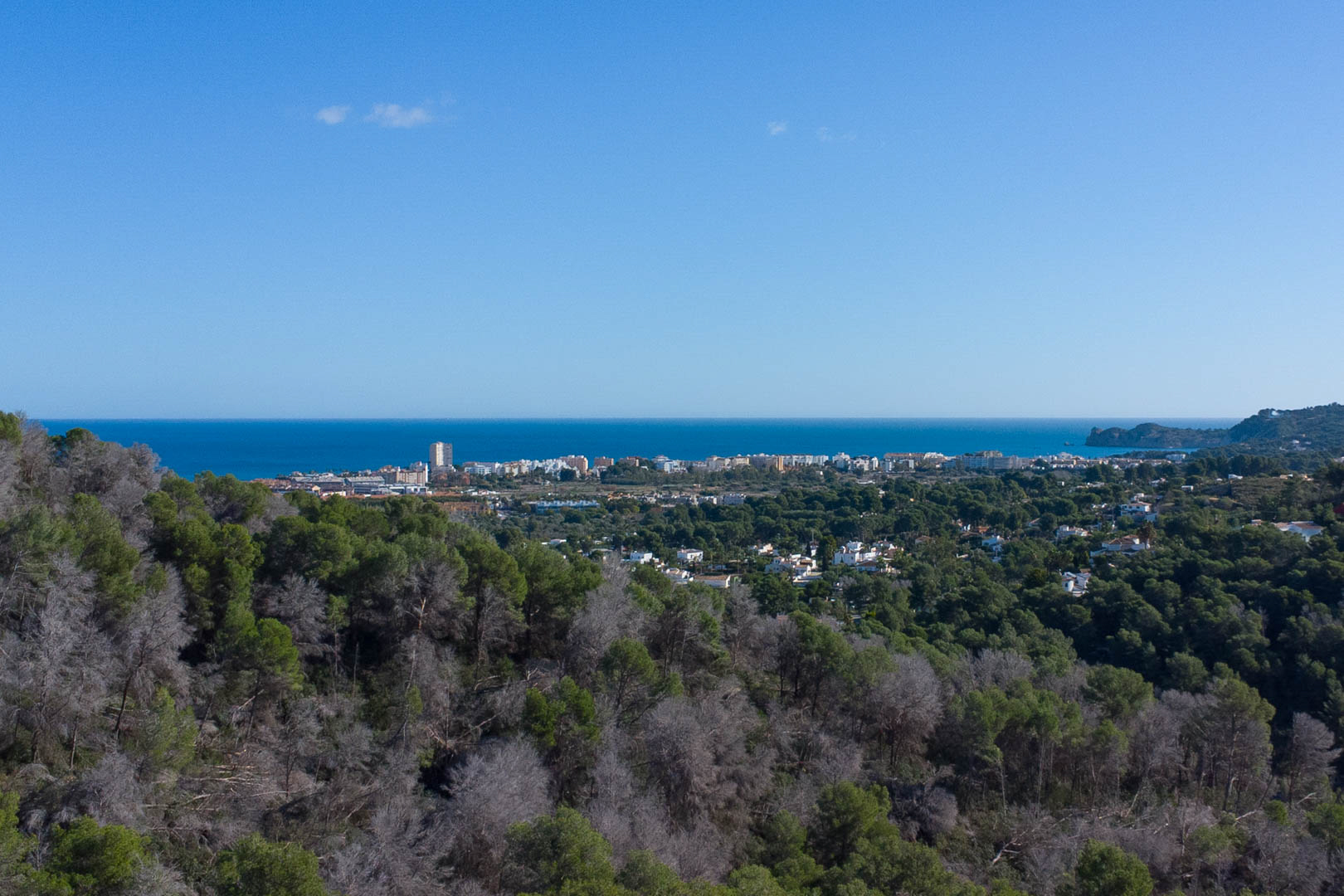 Panoramisch uitzicht vanaf het perceel over de Middellandse Zee en de kustplaats, omgeven door een dicht dennenbos onder een helderblauwe hemel.