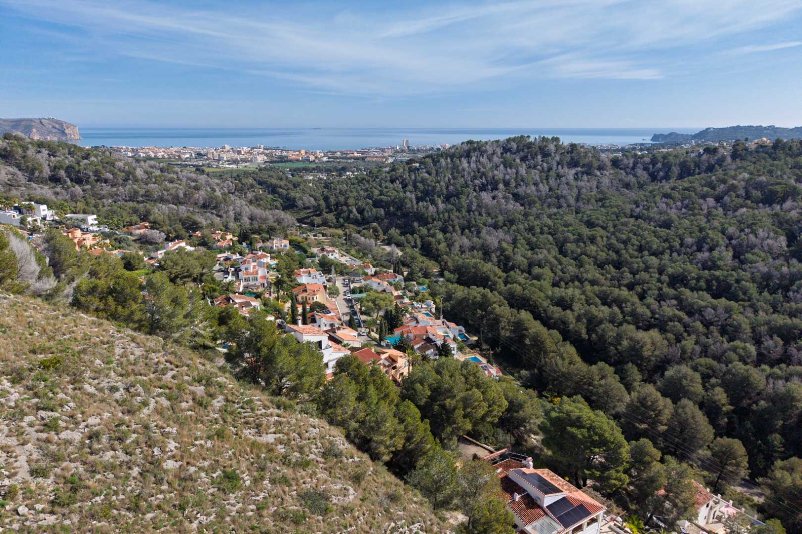 Panoramisch luchtfoto van de verhoogde ligging boven beboste heuvels, met vrij uitzicht op de Middellandse Zee en de kustlijn van Jávea in de verte.