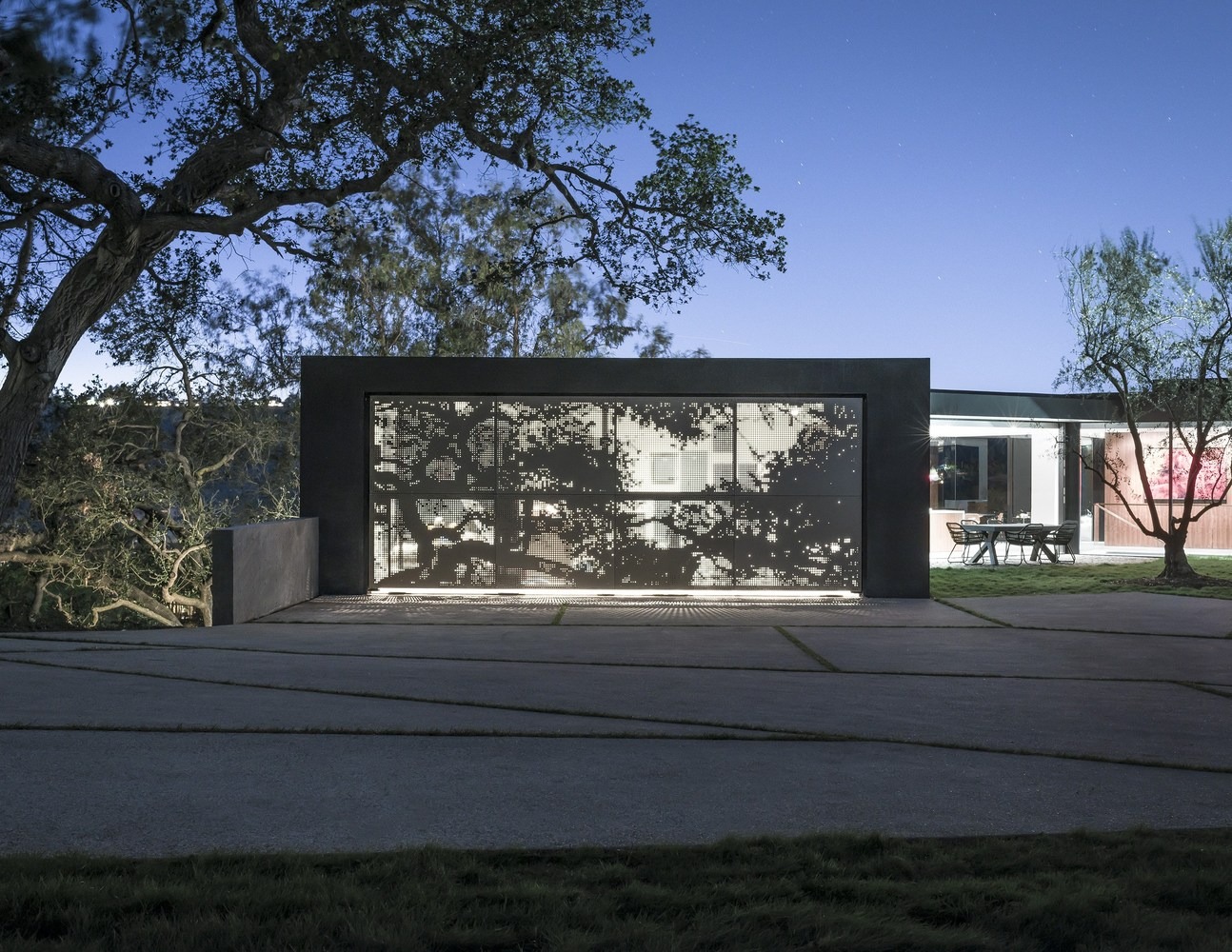 Moderne gevel met een verlicht geperforeerd metalen paneel met boommotieven, gecombineerd met geometrische bestrating en een aangrenzend buiteneetgedeelte.
