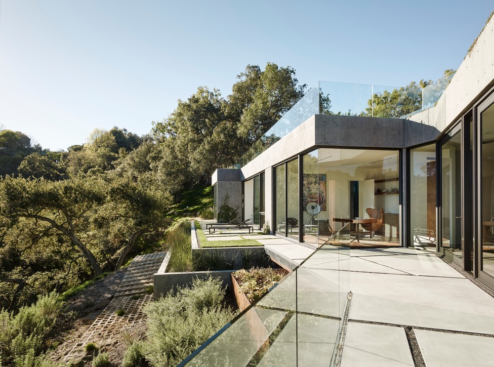 Moderne villa van beton met kamerhoge glazen wanden en een ruim terras met glazen balustrades en geometrische bestrating.