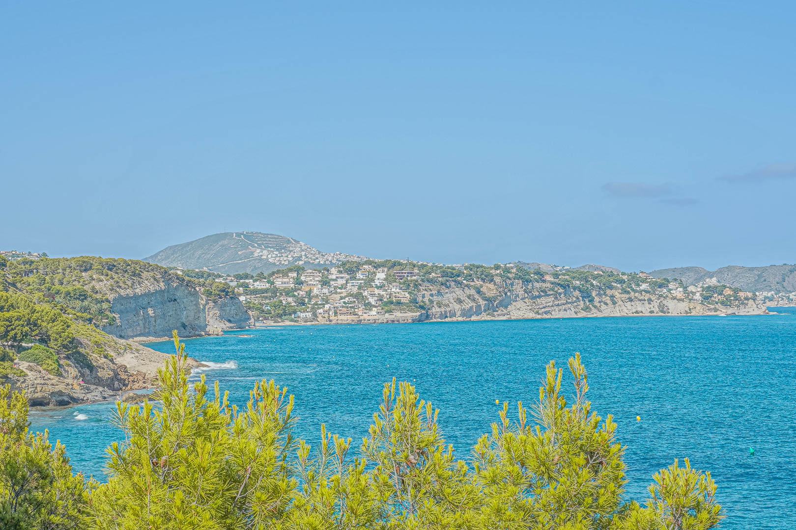 Panoramisch uitzicht op de kustlijn van Benissa met mediterrane pijnbomen op de voorgrond en residentiële kliffen boven de turquoise Middellandse Zee.