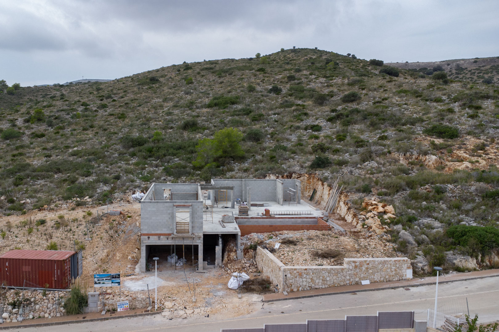Bouwfase van een villa in Pedreguer, met betonblokken structuren, een natuurstenen omheiningsmuur en integratie in de rotsachtige heuvelzijde.