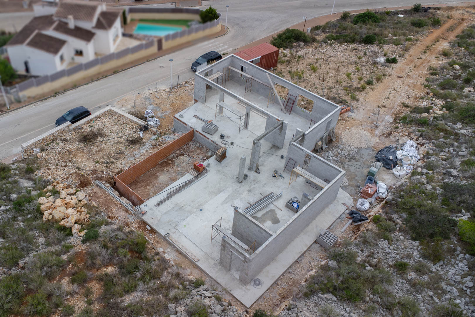 Luchtfoto van de betonnen fundering en de structuur van blokmuren van de villa in aanbouw, inclusief bouwmaterialen en steigers op het perceel.