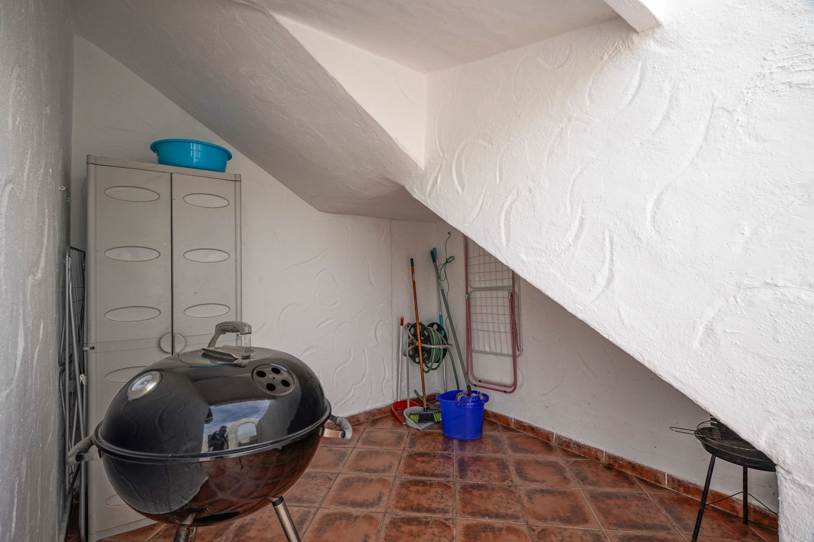 Buitenberging onder de trap met terracotta vloertegels, een kunststof opbergkast en ruimte voor een barbecue en onderhoudsmateriaal.