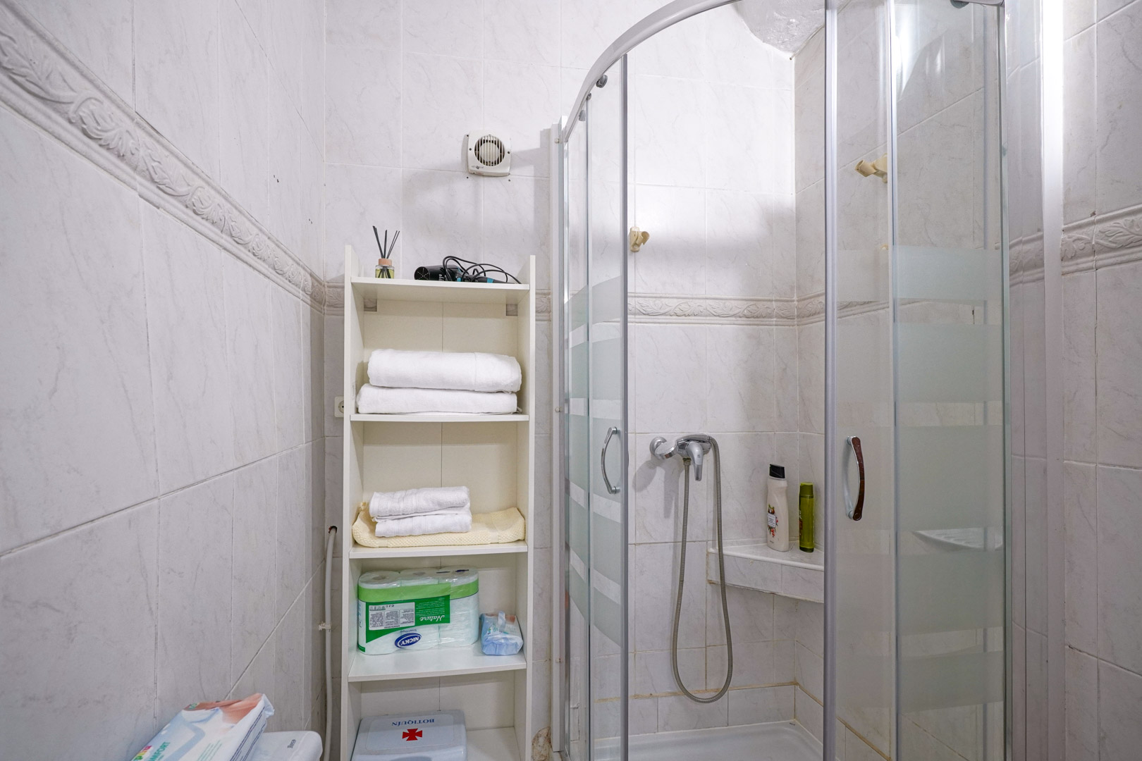 Functionele badkamer met een halfronde glazen douchecabine. De wanden zijn afgewerkt met witte tegels, een sierrand en een wit opbergrek voor handdoeken.