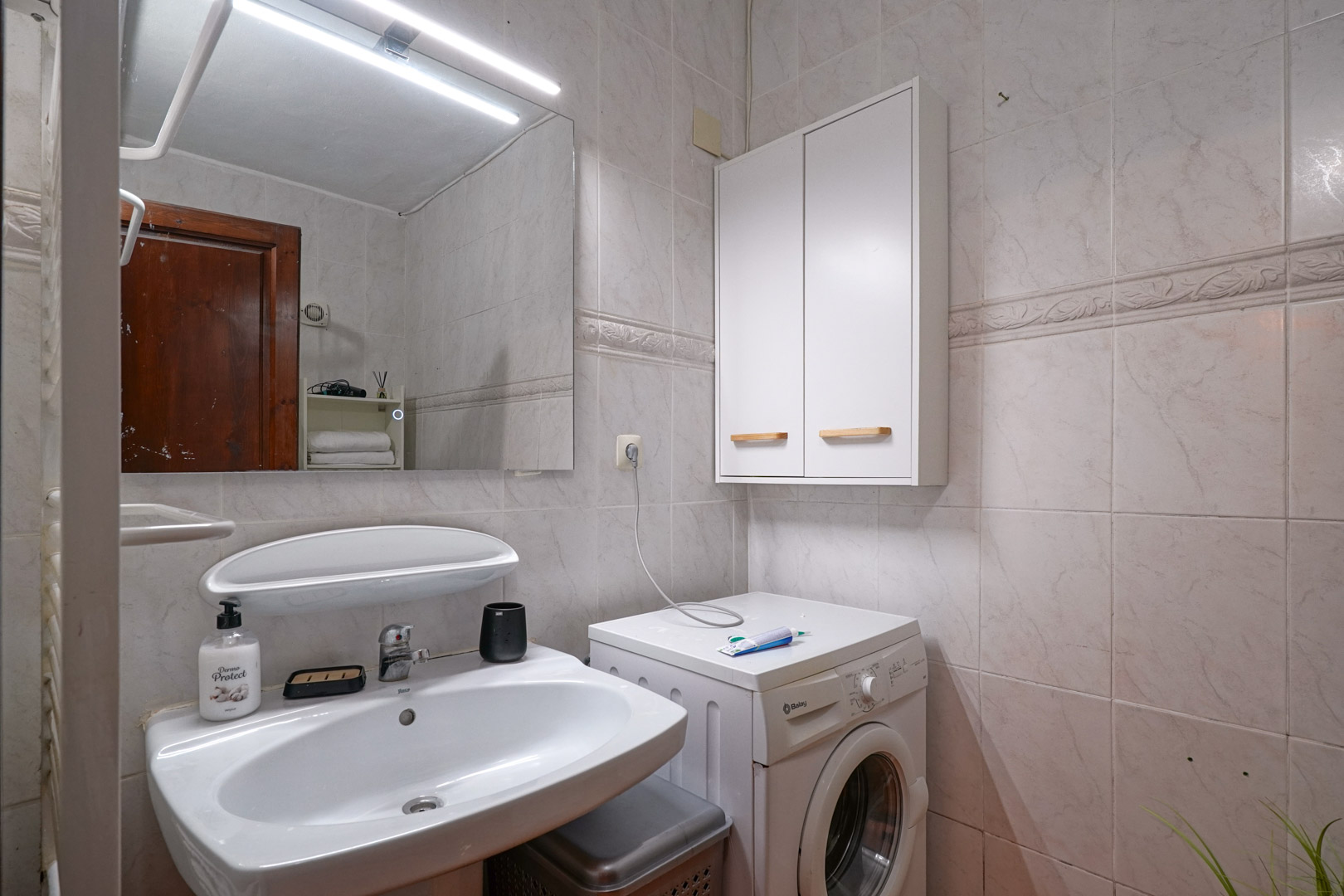 Functionele badkamer met keramische wastafel, spiegel met LED-verlichting en geïntegreerde wasruimte met wasmachine en wandkast tegen licht betegelde muren.