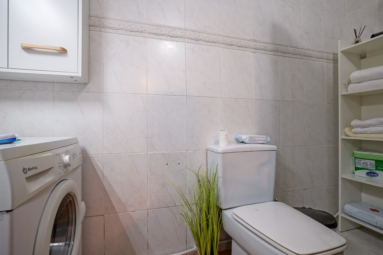 Functionele badkamer met wasruimte, voorzien van een Balay wasmachine, opbergplanken en betegelde wanden met een decoratieve rand.