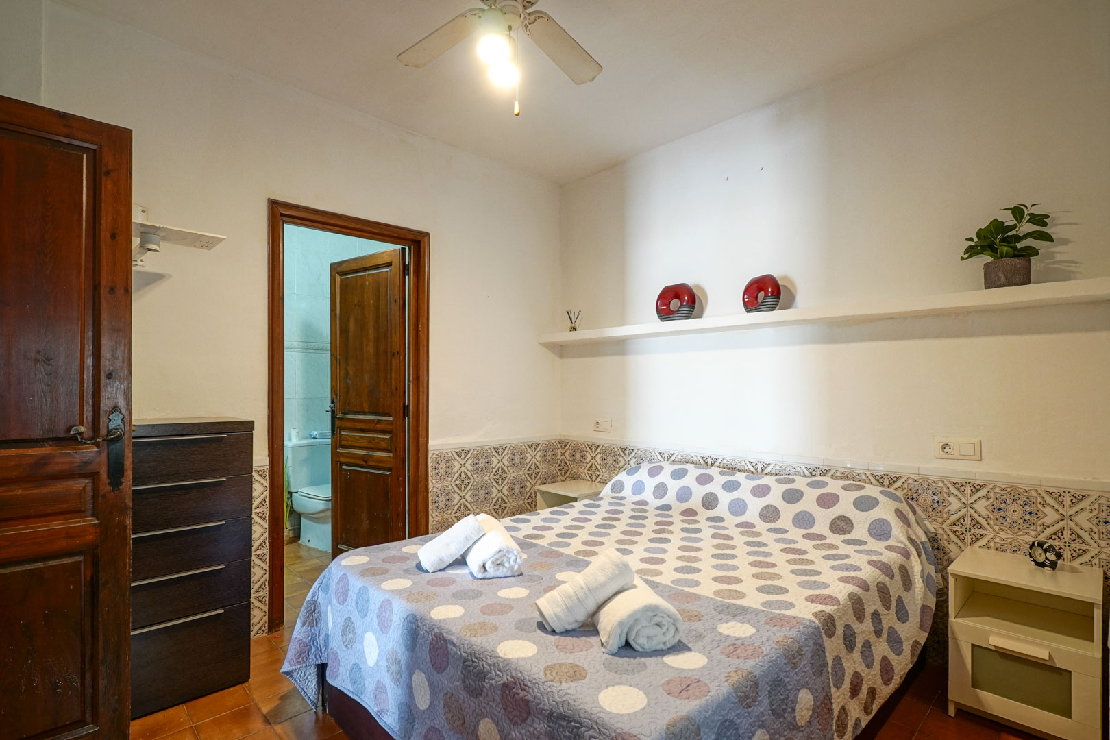Hoofdslaapkamer suite met decoratieve tegellambrisering, een plafondventilator en directe toegang tot de aangrenzende badkamer via een houten deur.