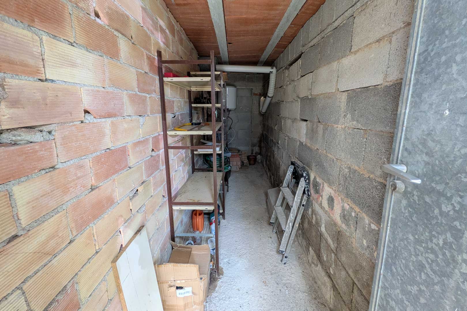 Onafhankelijke berging met wanden van baksteen en betonblokken, voorzien van metalen rekken en een elektrische boiler achterin.