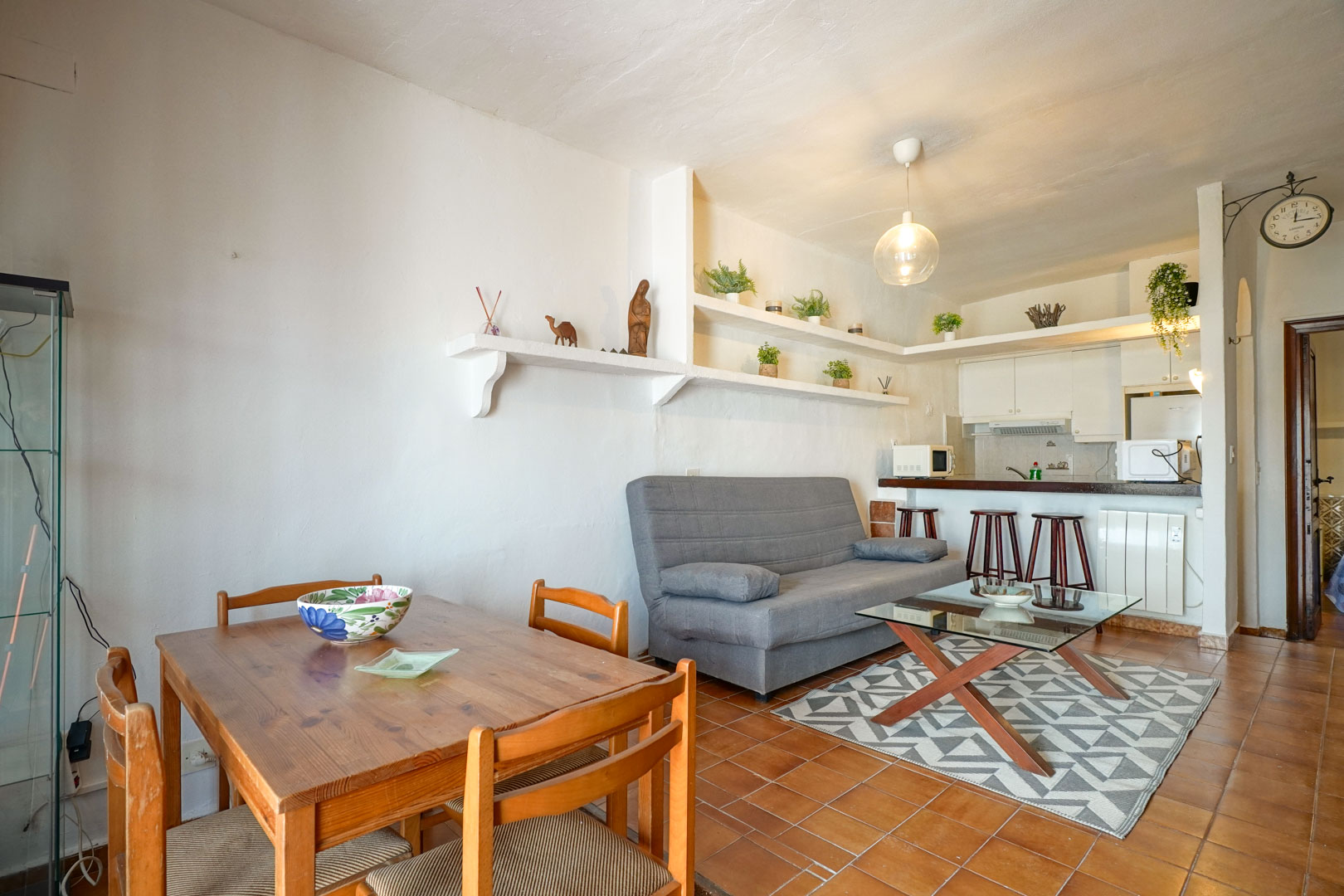 Open woon- en eetkamer met terracotta vloertegels, ingebouwde witte wandplanken en een halfopen keuken met ontbijtbar en houten krukken.