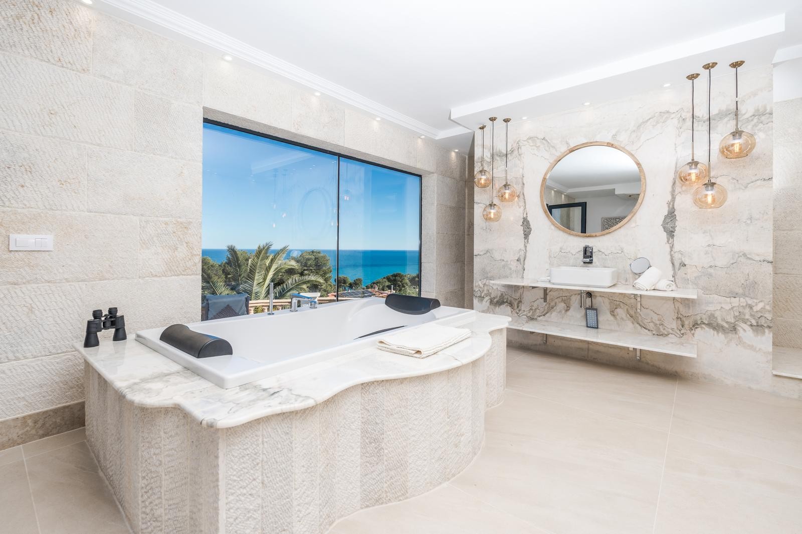 Badkamer van de master suite met een in marmer gevat ligbad bij het raam met zeezicht, natuurstenen wanden en een wastafelmeubel met marmeren bladen en designverlichting.