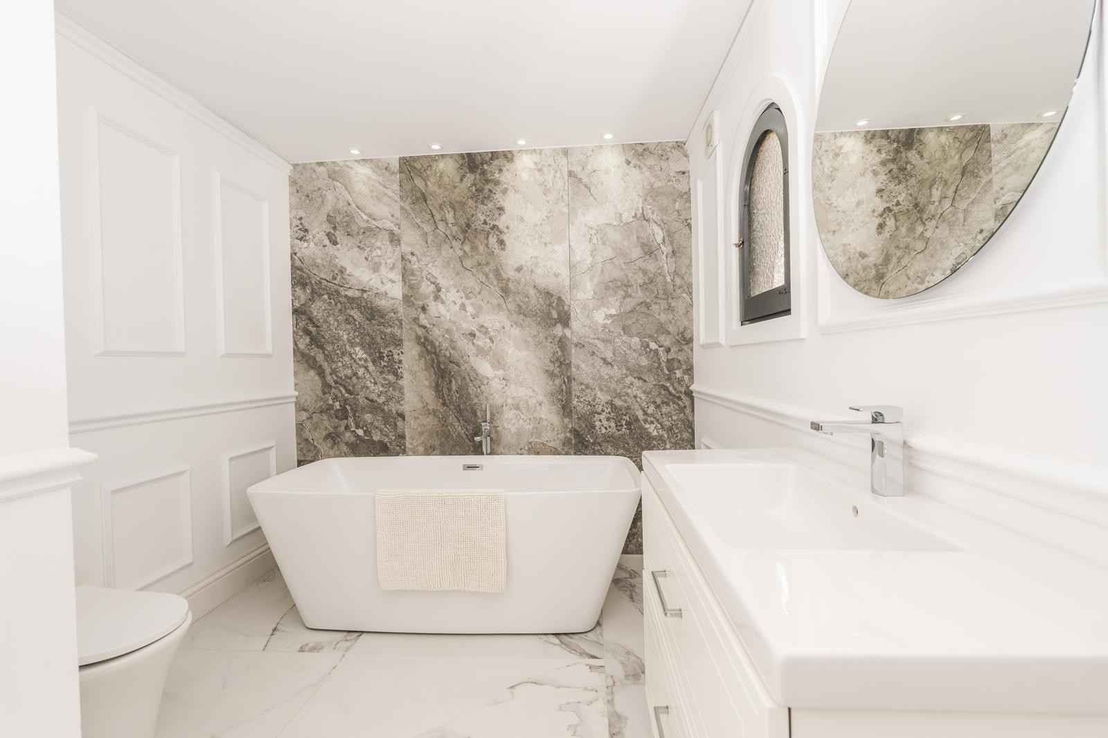 Badkamer van de master suite met vrijstaand bad, marmeren accentmuur en sierlijsten. Voorzien van een wit wastafelmeubel, ronde spiegel en inbouwspots.