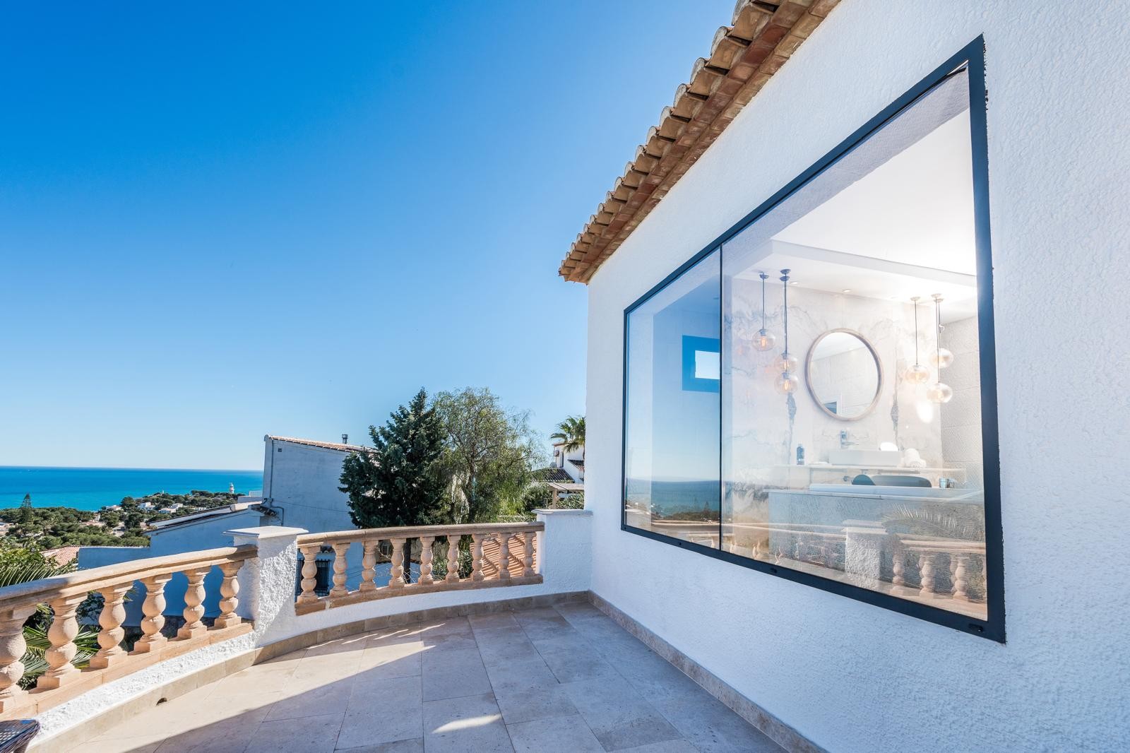 Bovenste terras met stenen balustrade en zeezicht, grenzend aan een groot raam dat uitkijkt op een moderne badkamer met marmeren wanden en designverlichting.