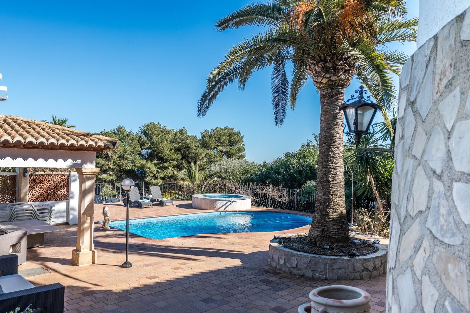 Buitenterras met een niervormig zwembad, ronde jacuzzi en een grote palmboom. Voorzien van een overdekte veranda met stenen zuilen en authentieke terracotta tegels.