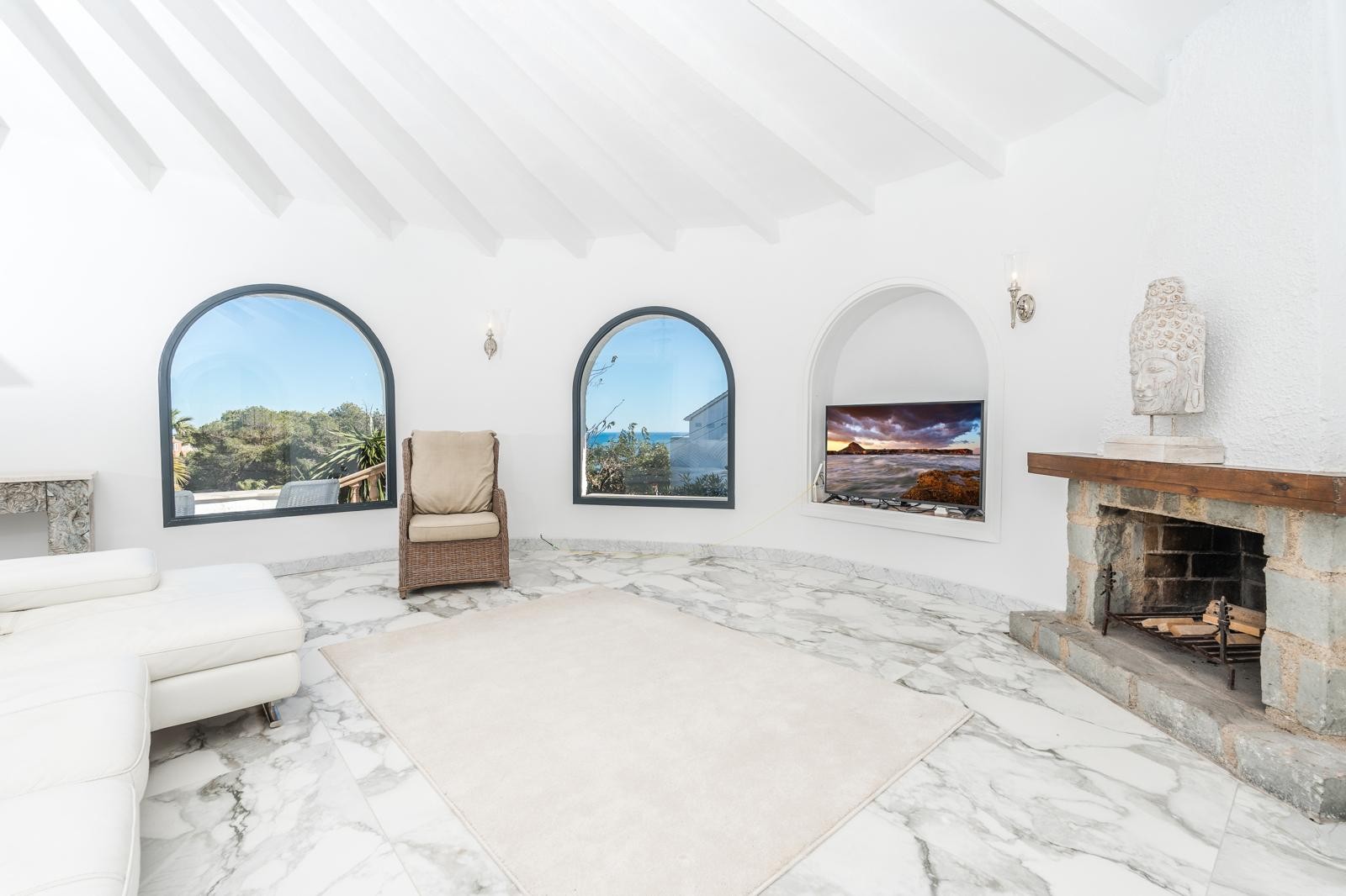 Cirkelvormige woonkamer met wit balkenplafond, marmeren vloer en een stenen open haard. Boogvensters zorgen voor natuurlijk licht en uitzicht op de mediterrane omgeving.