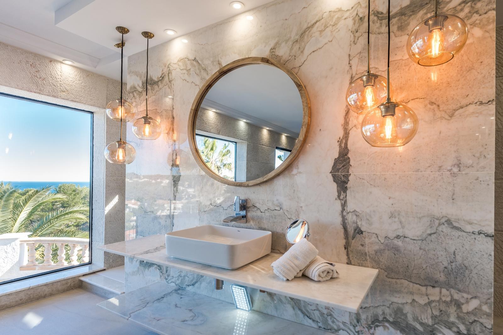 Designbadkamer met geaderde marmeren wand en werkblad, een ronde houten spiegel en glazen hanglampen met uitzicht op de Middellandse Zee.