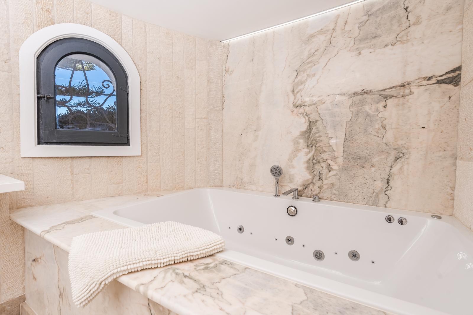 Ingebouwd hydromassagebad met marmeren afwerking en wanden van natuursteen. Voorzien van een karakteristiek boogvenster en subtiele LED-verlichting aan het plafond.