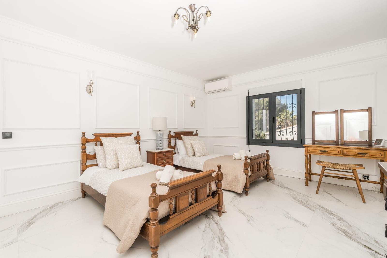 Lichte slaapkamer met twee houten eenpersoonsbedden, marmeren vloer met grijze aders en klassieke wandlijsten. Voorzien van een houten kaptafel en split-airconditioning.