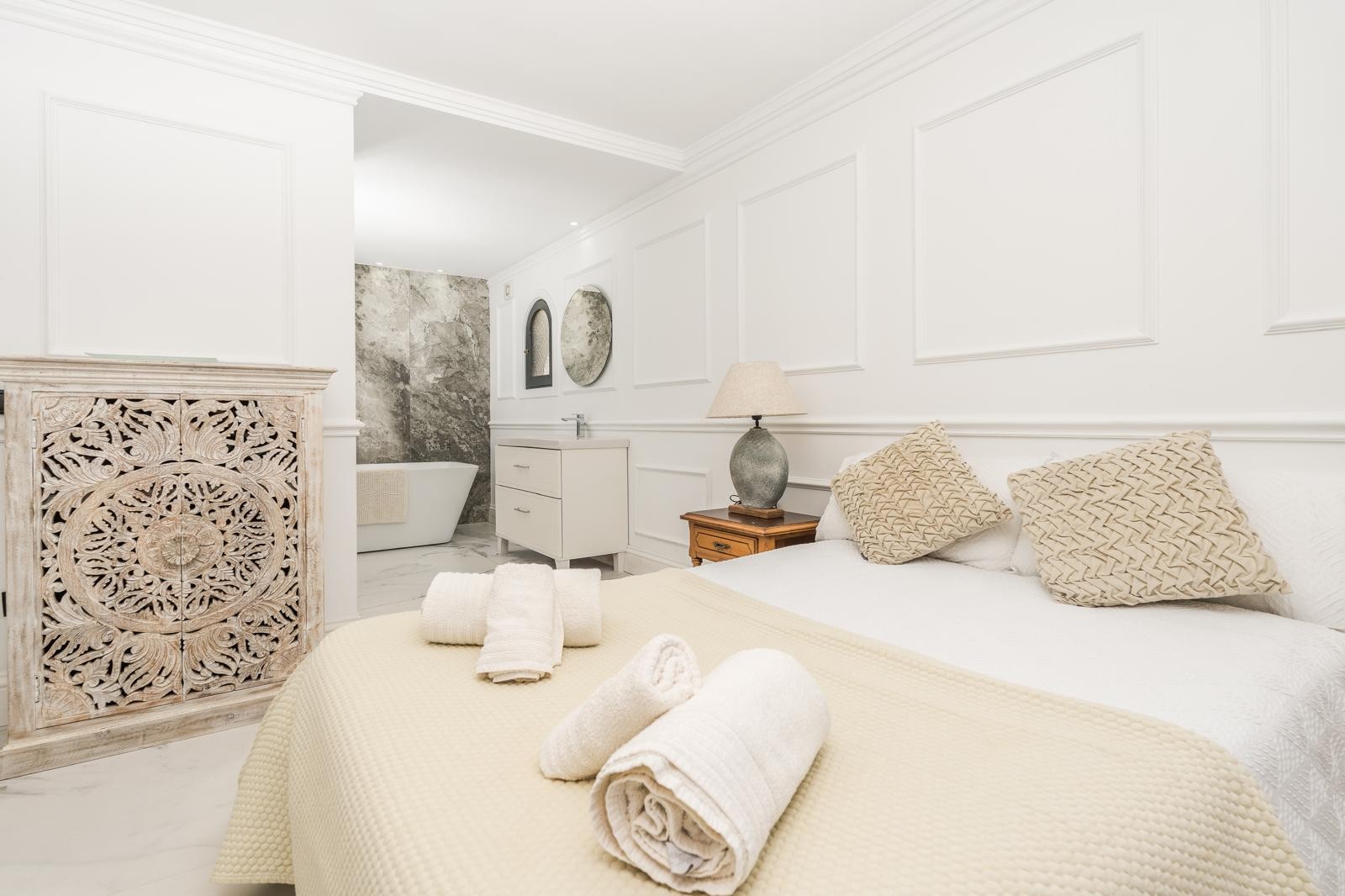 Master suite met open badkamerconcept, voorzien van een vrijstaand bad tegen een grijze marmeren wand, klassieke wandlijsten en een handgesneden houten kast.