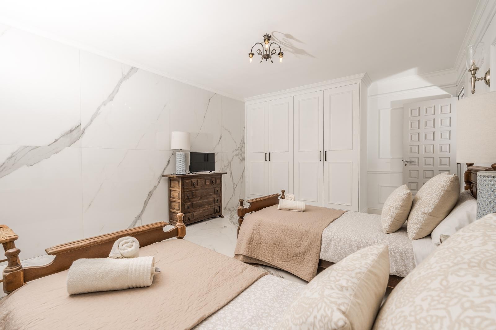 Slaapkamer met twee houten eenpersoonsbedden, een wand met marmerlook en witte inbouwkasten. Een rustieke ladekast vult de lichte, moderne afwerking aan.
