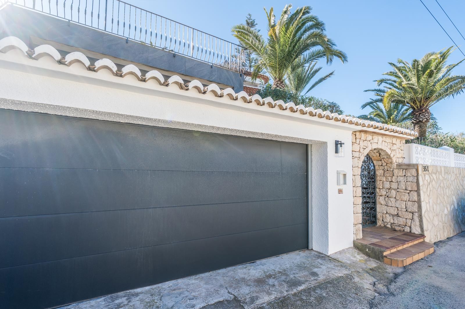 Vooraanzicht met een dubbele garage voorzien van een antracietkleurige automatische deur, een natuurstenen muur met boogingang en mediterrane palmen.