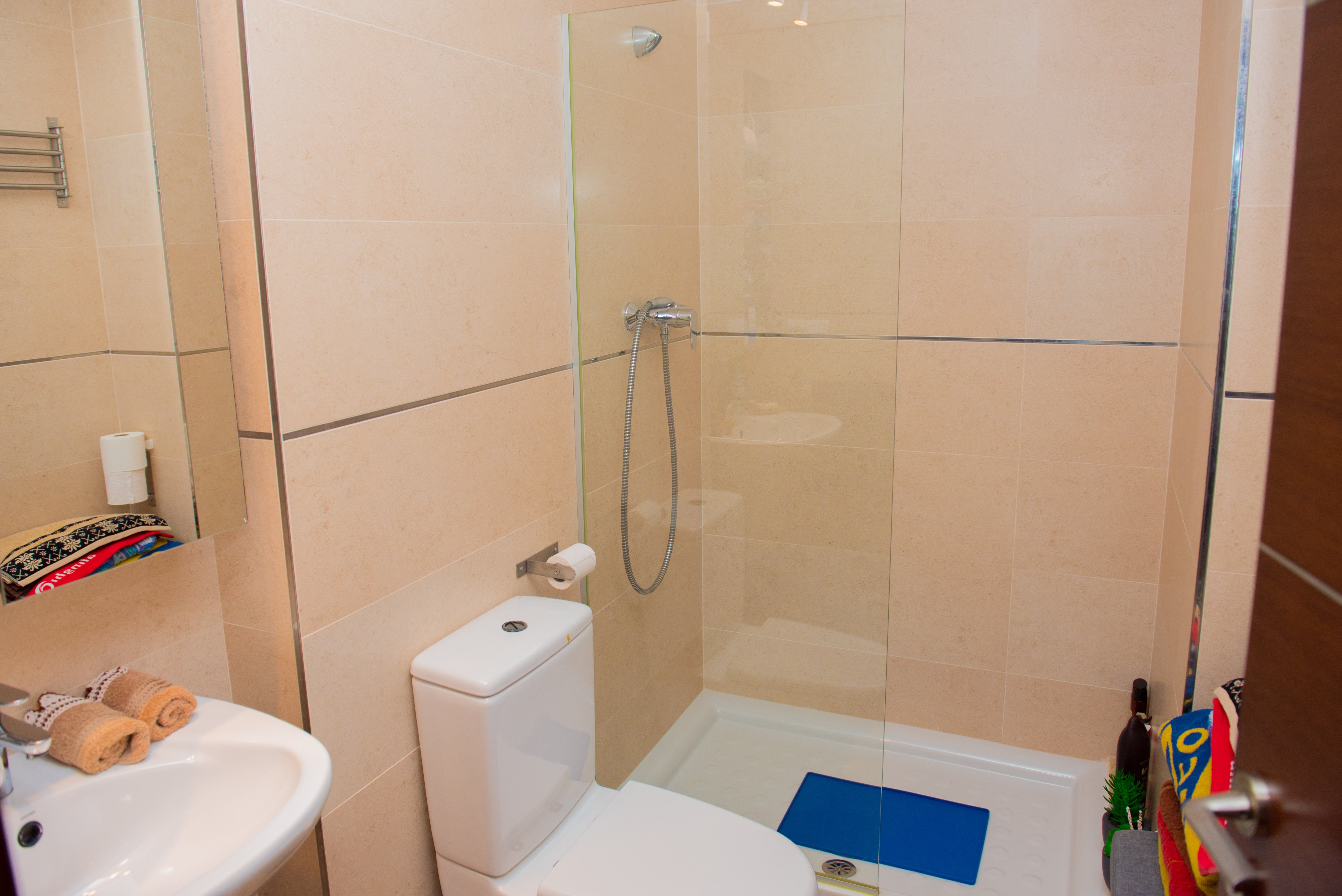 Moderne badkamer met inloopdouche, glazen wand, wit toilet en crèmekleurige wandtegels afgewerkt met chromen sierlijsten.
