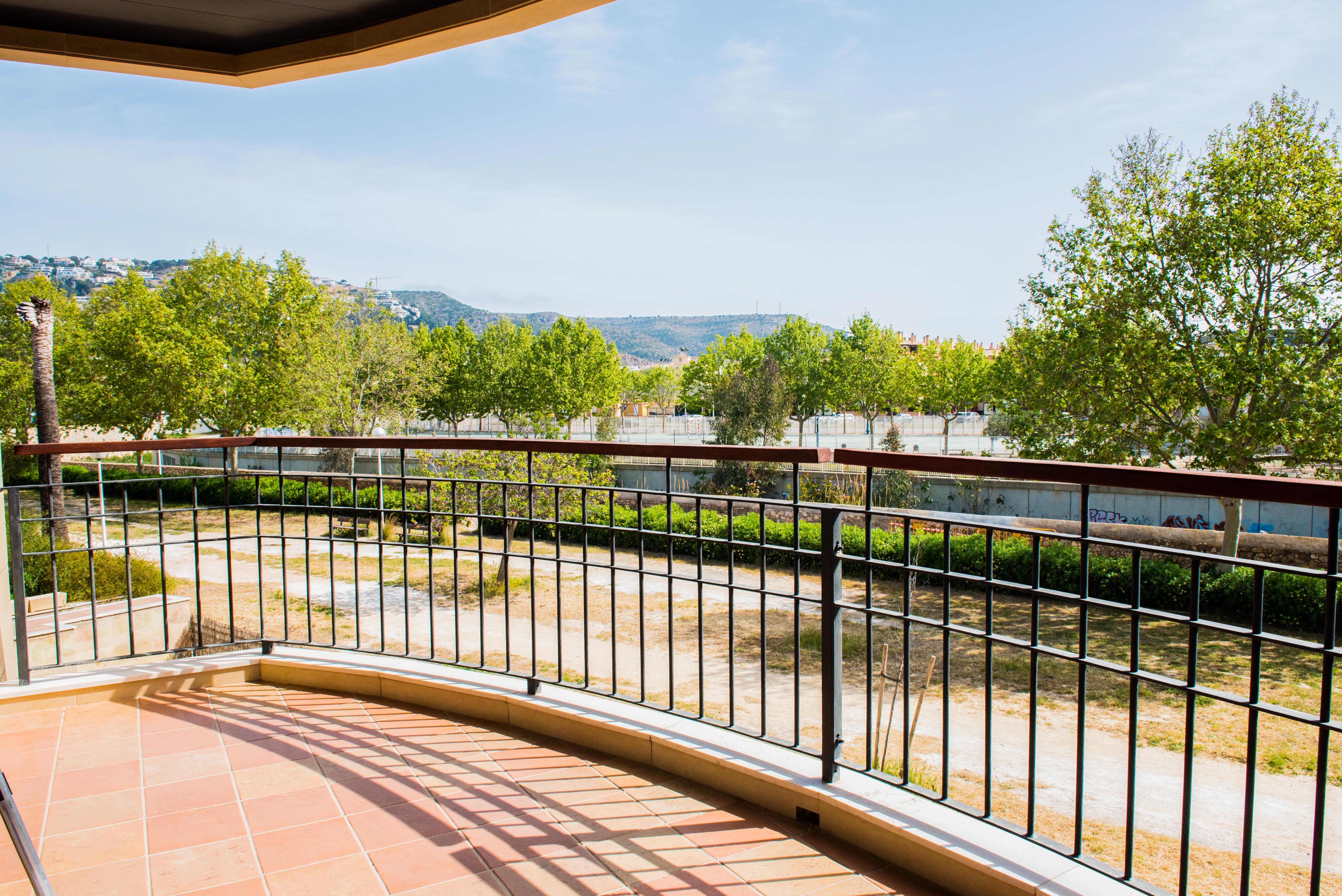 Overdekt terras met terracotta tegelvloer en smeedijzeren balustrade met houten handgreep, uitkijkend over een groen park en de bergen.