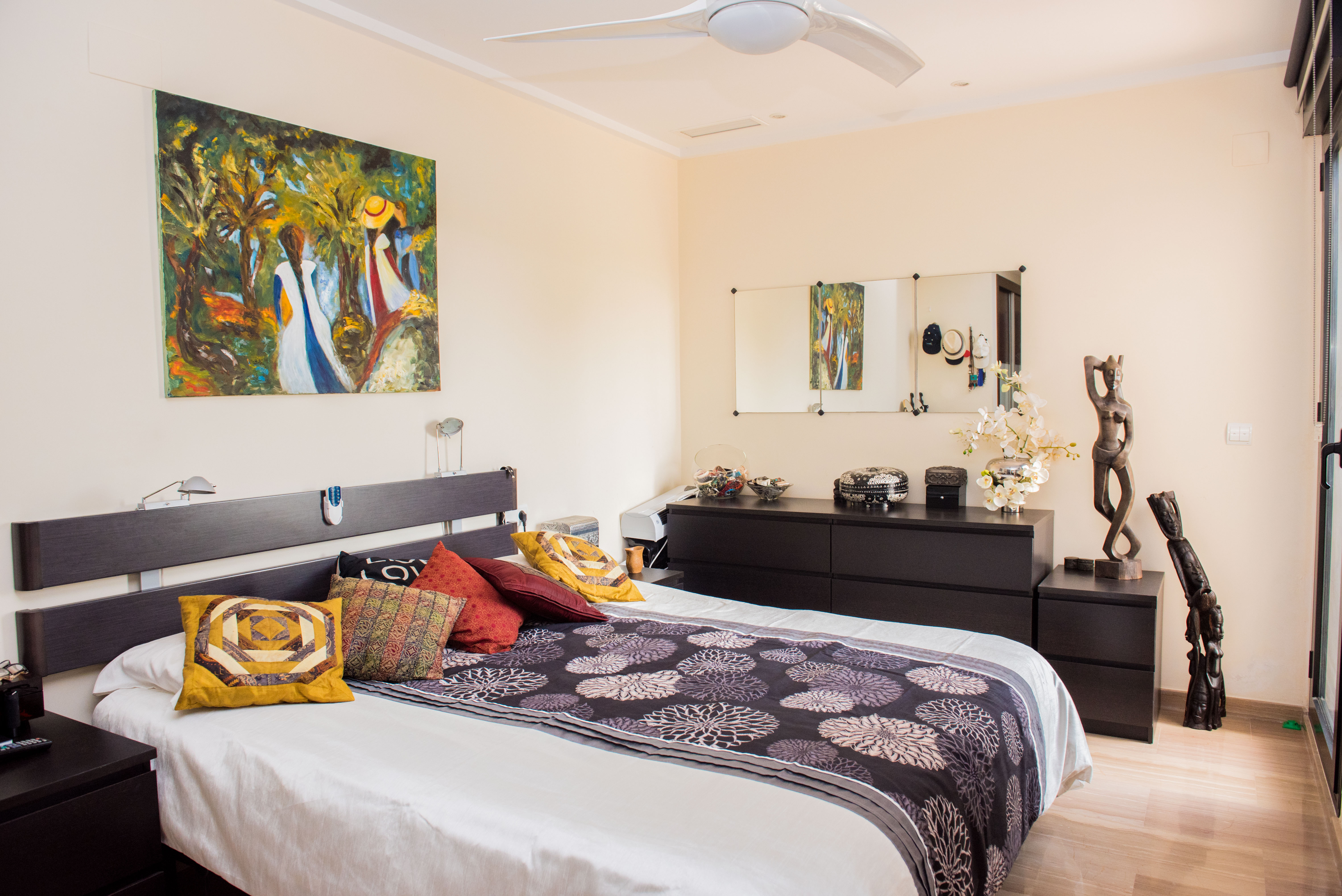 Slaapkamer met donkerhouten hoofdbord, bijpassende ladekast en plafondventilator. Voorzien van kleurrijke kunst, decoratieve beelden en een lichte vloer.
