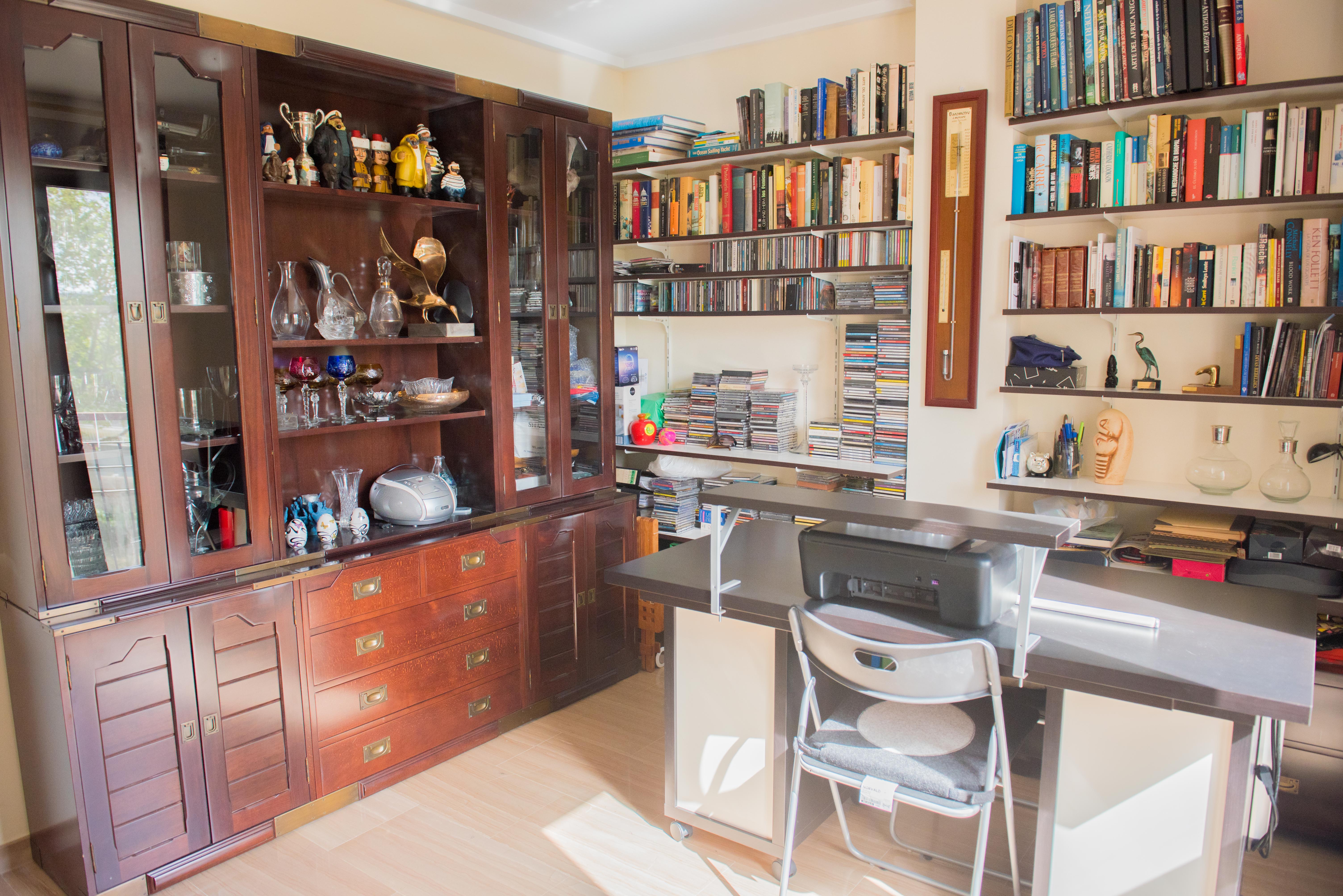 Werkkamer met een grote donkerhouten vitrinekast, uitgebreide wandplanken vol boeken en een functioneel bureau met lichte tegelvloer en natuurlijk licht.